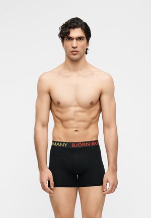 Model masculin cu păr creț închis la culoare, purtând boxeri negri Björn Borg cu text roșu și galben pe talie, stând pe un fundal alb.