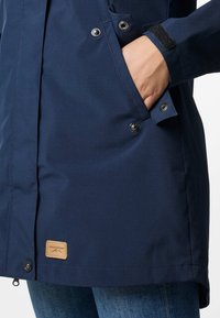 Giacca blu navy in tessuto morbido, con una mano in tasca, chiusure a scatto e un'etichetta in pelle con logo sull'orlo inferiore.