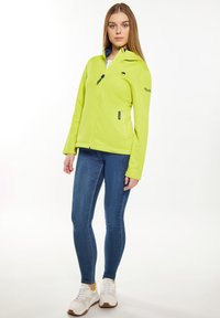 Giacca in pile verde lime brillante con zip frontale, due tasche laterali e logo sul petto. Abbinata a jeans skinny blu e sneaker bianche.