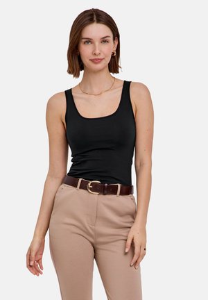 Vrouw met kort bruin haar, gekleed in een zwarte mouwloze top, beige broek met een bruine riem en gouden sieraden, staand tegen een witte achtergrond.