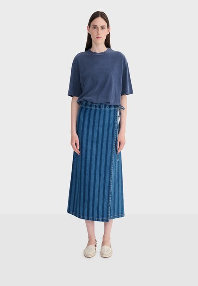 Woman standing facing forward wearing a loose navy blue t-shirt, long blue striped denim skirt with tie detail, and white slip-on shoes.