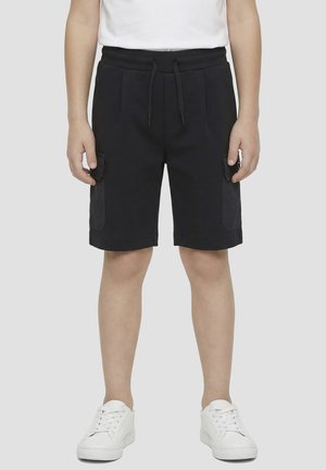 Niño con pantalones cortos cargo negros hasta la rodilla con cintura de cordón y zapatillas blancas sobre un fondo liso.