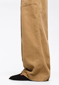 Pantalones de pierna ancha beige texturizados con un patrón sutil, combinados con zapatos negros de punta afilada que presentan acentos rojos en los laterales.