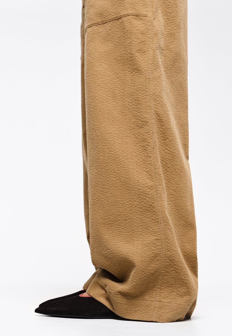 Pantalones de pierna ancha beige texturizados con un patrón sutil, combinados con zapatos negros de punta afilada que presentan acentos rojos en los laterales.