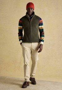 Zielona polarowa kamizelka nałożona na koszulę z długim rękawem w kolorowe paski, kremowe spodnie i brązowe sznurowane buty, do tego czerwona dziergana czapka.