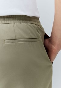 Olive green pants featuring a smooth texture, elastic waistband, and a single back pocket with a sleek opening.