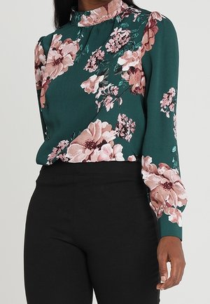 Femme portant un chemisier vert à motif floral rose et col montant, associé à un pantalon noir taille haute.