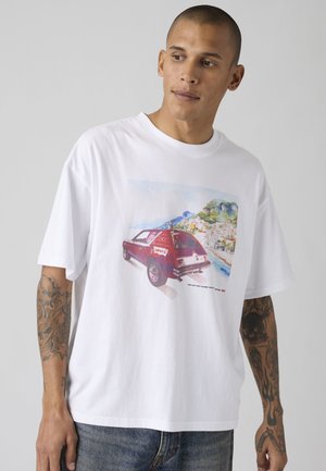 Homme avec tatouages portant un t-shirt blanc avec un graphisme de voiture rouge et une ville côtière pittoresque, associé à un jean bleu, sur un fond uni.