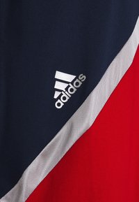 Maillot de sport bleu marine, rouge et argent avec un design en blocs de couleur diagonaux. Présente un logo Adidas blanc dans le coin inférieur gauche. Matière semblable au coton.