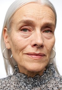 Gros plan sur une personne aux cheveux argentés, portant un chemisier floral noir et blanc aux bords volants et de grandes boucles d'oreilles en argent. Sa peau présente des ridules.
