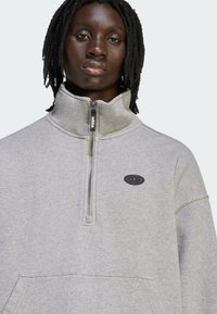Felpa grigia con cerniera in pile, collo alto, tasca frontale e toppa logo nera. Presenta una chiusura con cerniera e una texture morbida.