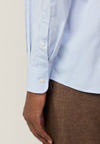 Camisa de vestir a rayas azul claro y blanca con puños abotonados, de tela texturizada visible, combinada con pantalones de espiga marrón.