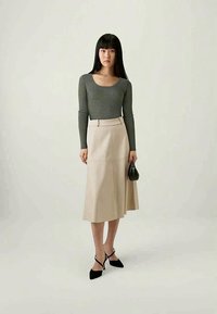 Beige A-linjekjol med bälte i midjan, kombinerad med en åtsittande grå långärmad topp. Modellen håller i en liten svart handväska och har svarta klackskor.