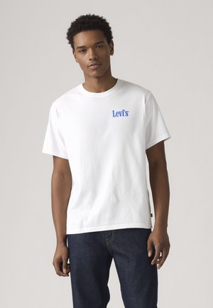 Νέος άνδρας που φοράει λευκό T-shirt Levi’s και σκούρο μπλε τζιν, στέκεται μπροστά σε ουδέτερο φόντο.