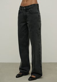 Aukštos juosmens juodos džinsinės kelnės su tiesiomis kojomis, turinčios priekinės kišenes, subtilius siūlus ir lygią tekstūrą.