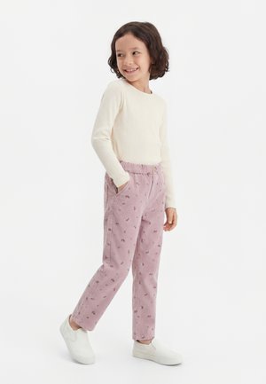 Enfant portant un haut crème à manches longues côtelé, un pantalon rose à fleurs et des chaussures blanches à enfiler, debout les mains dans les poches et souriant.