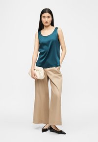 Mujer con blusa sin mangas de satén color verde azulado, pantalones beige de pierna ancha, zapatos planos negros puntiagudos, sosteniendo un clutch blanco, de pie frente a un fondo liso.