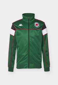 Chaqueta deportiva verde hecha de tejido suave. Presenta un cuello alto, cremallera completa, paneles en blanco y negro, acentos rojos y un logo de estrella.