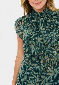 Frau in bluse mit grünen Paisleymuster, Rüschen, hochgeschlossenem Kragen und kurzen Ärmeln.
