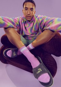 Kolorowy hoodie tie-dye w różowym i zielonym kolorze, spodenki w wzory, różowe skarpetki do kostek w paski oraz szare klapki, wszystko na brązowym pluszowym fotelu.