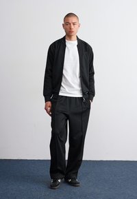 Costum sport negru format dintr-o jachetă bomber cu fermoar și pantaloni largi, asortat cu un tricou alb și adidași negri. Material neted.