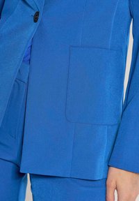 Donkerblauwe colbert met een enkele knoopsluiting, met gesneden revers en een paspelzak met zichtbare stiksels. Gladde textuur.