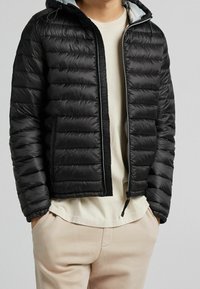 Osoba v černé prošívané bundě s kapucí, pod kterou má béžovou košili a béžové kalhoty, ruce v kapse, na jednobarevném pozadí.