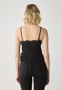 Svart camisole med blonder trim ved halsen og kanten. Justerbare tynne stropper, laget av et glatt stoff, sammen med svarte jeans.