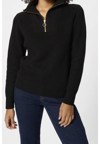 Schwarzer Pullover mit Reißverschluss am Kragen, gerippter Struktur und goldfarbenem Reißverschlussakzent, mit langen Ärmeln und figurbetontem Schnitt. Kombiniert mit dunklen Jeans.