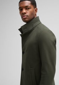 Olijfgroen coat met een hoge kraag, dubbele knopen, zijzakken en een gestructureerde stof. Het getailleerde model is zichtbaar op het model.