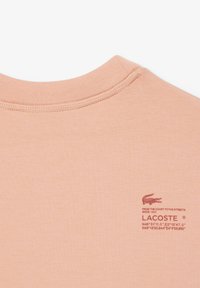 Ferskenfarget bomulls-t-skjorte med ribbet rund hals og en liten rød Lacoste-logo og tekst nederst til høyre. Glatt tekstur.