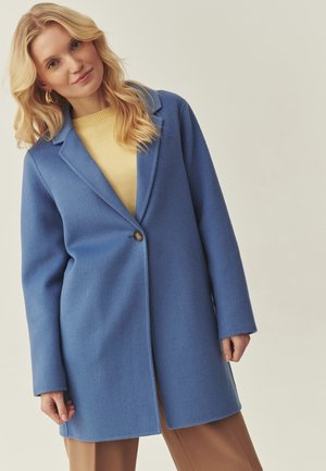 Cappotto blu in lana con colletto a risvolto, chiusura con un solo bottone e maniche lunghe, indossato sopra un maglione giallo e pantaloni marroni.