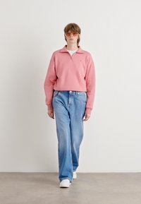Pullover in pile rosa con colletto a zip, abbinato a jeans larghi blu chiaro e sneakers bianche, su uno sfondo neutro. Textures chiare e morbide.