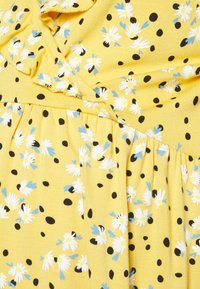 Tecido amarelo com margaridas brancas e detalhes em azul, sobreposto com bolinhas pretas. Apresenta um decote com babados e detalhe de cintura franzida.