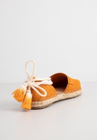 Espadrille orange à enfiler avec semelle en jute tressée et nœud décoratif en corde crème avec pompons orange et blanc.