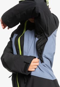 Giacca nera e blu chiaro con dettagli verde fluo sulla cerniera. Presenta una finitura testurizzata, polsini regolabili e una fodera interna in rete.
