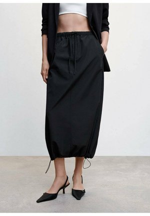 Femme portant une jupe midi noire à cordon, un blazer noir, un top blanc court et des escarpins noirs pointus à bride arrière sur un sol gris.