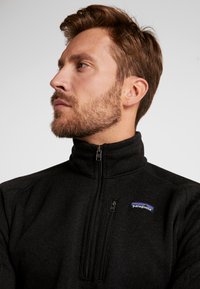 Schwarzer Fleece-Pullover mit hohem Kragen und Reißverschluss am Hals. Verfügt über ein blau-weißes Patagonia-Logo auf der linken Brust. Strukturierten Stoff.