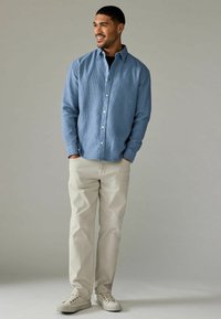 Cămașă bleu deschis, cu un material texturat, pantaloni bej drepti și adidași crem. Modelul stă cu mâinile în buzunare.