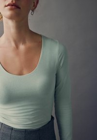 Lys grøn langærmet top med rund halsudskæring, tætsiddende tekstur og glat stof; parret med en mørkegrå plisseret nederdel.
