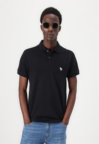 Črna polo majica iz bombaža, z ovratnikom in kratkimi rokavi. Izvezen logotip v beli barvi na levi strani prsnega koša. Nosi se z modrimi kavbojkami.