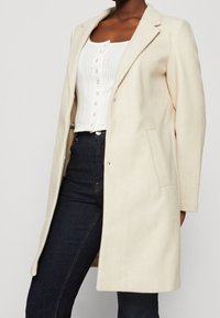 Crème kleurige jas gemaakt van zachte stof, met een opstaand kraag, schoudernaden en twee voorkeurszakken, gedragen over een witte top.