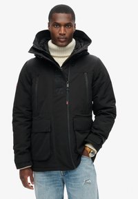 Chaqueta de plumón negra con cuello alto, que presenta un cierre frontal, dos bolsillos laterales y un detalle de logo. Llevada sobre un suéter de cuello alto color crema.
