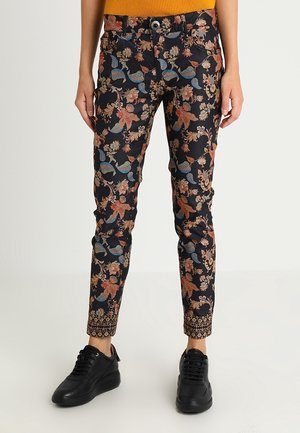 Pantalon noir slim fit avec motifs floraux et feuillages multicolores, poignets décoratifs à motifs, porté avec des baskets noires et un haut orange.