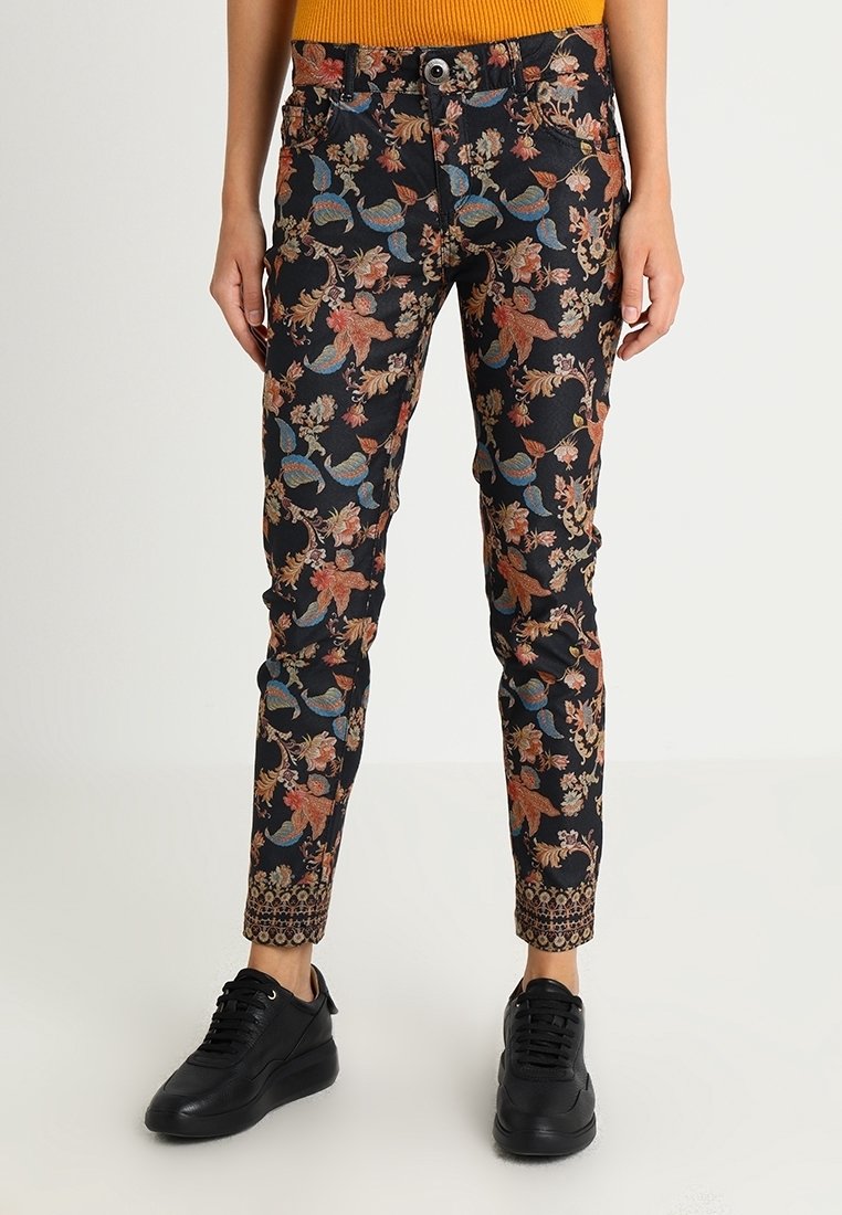 Pantalon noir slim fit avec motifs floraux et feuillages multicolores, poignets décoratifs à motifs, porté avec des baskets noires et un haut orange.