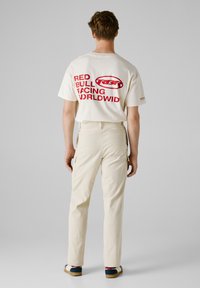 Witte t-shirt met een rode opdruk met de tekst "RED BULL FACING WORLDWID" op de rug. Beige broek met zakken, gedragen met blauw-witte sneakers.