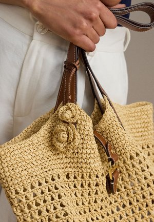 Ręka trzyma tkany słomiany torebkę z brązowymi skórzanymi uchwytami i kwiatowymi zdobieniami, noszona z białymi spodniami.