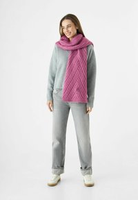 Vrouw in een grijze trui, lichtgrijze jeans, witte sneakers en een gestructureerde roze sjaal om haar nek, die glimlacht en naar links kijkt.