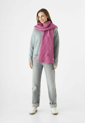 Vrouw in een grijze trui, lichtgrijze jeans, witte sneakers en een gestructureerde roze sjaal om haar nek, die glimlacht en naar links kijkt.