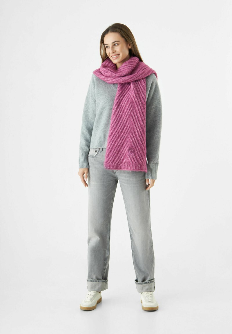 Vrouw in een grijze trui, lichtgrijze jeans, witte sneakers en een gestructureerde roze sjaal om haar nek, die glimlacht en naar links kijkt.